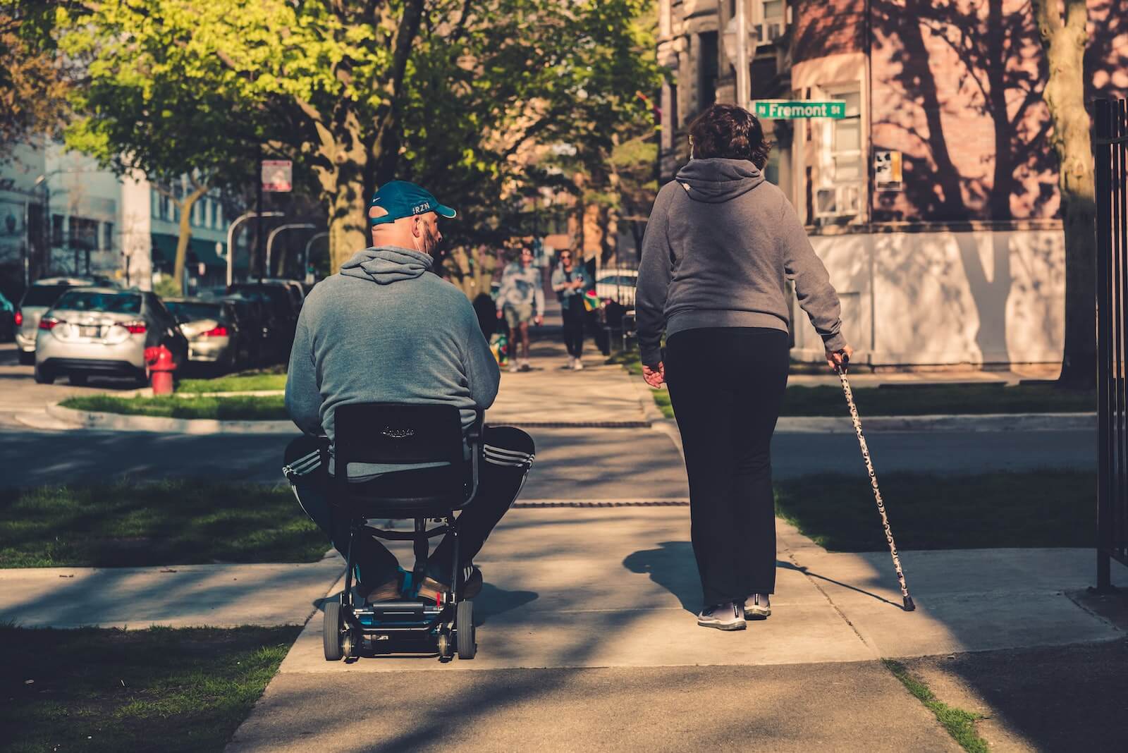 man in wheelchair and woman with cane walking on sidewalk